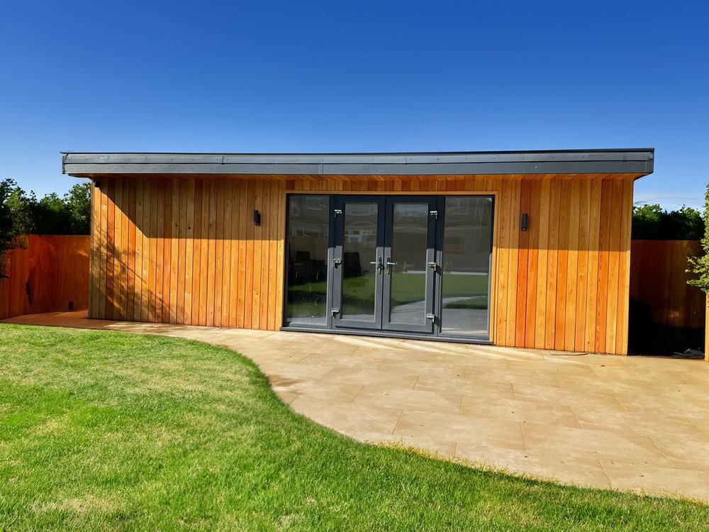 Vertical wood cladded garden room, anthracite patio doors with up and down lights. Paved area leading to the entrance and grass surrounds.